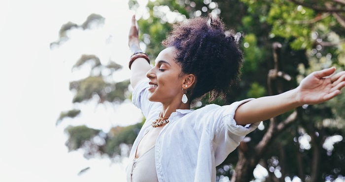 A young Black woman in a white shirt looking peaceful, standing in the sun with her arms outstretched and prioritizing her mental health while preparing for the CPA Exam.