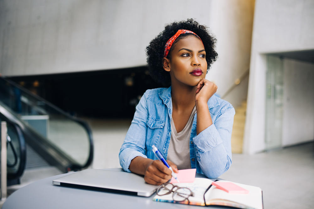 Focused young African-American female deep in thought while working at a table in a modern office building lobby.