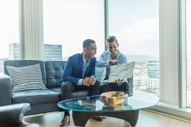 two men sitting on a couch and smiling at a laptop