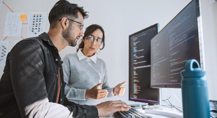 Young man with beard and glasses sitting at a computer with a young woman with long dark hair and glasses standing beside him. They are using AI in auditing